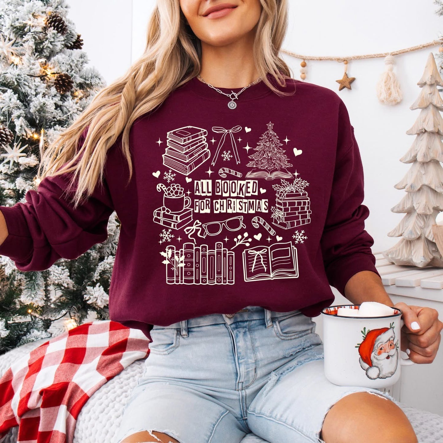 sweatshirt with a white holiday book-themed design and the phrase βAll Booked for Christmas,β styled flat lay with festive decor including pine branches, red berries, and cozy reindeer slippers.