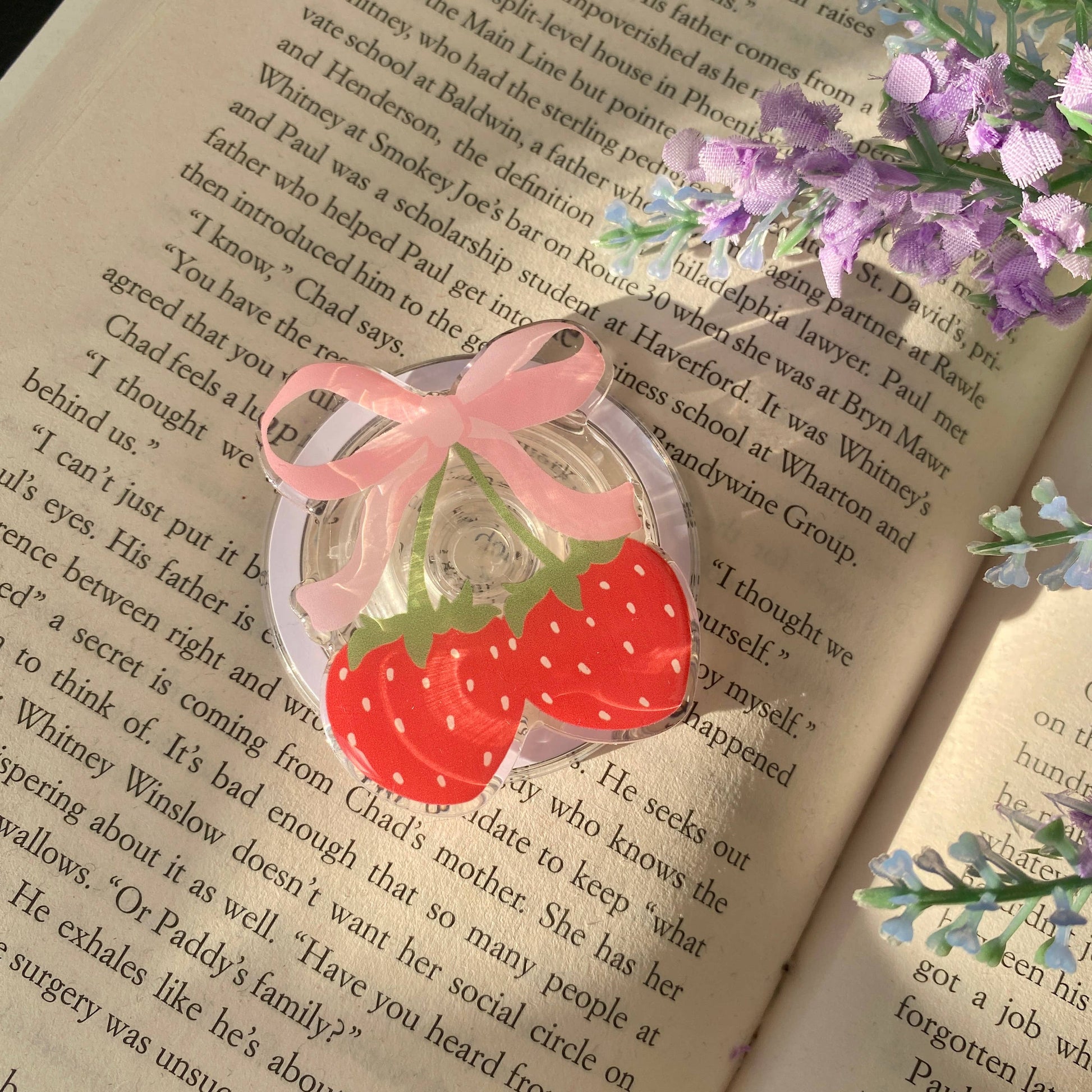 Strawberry-shaped hair tie with pink bow on an open book with purple flowers.