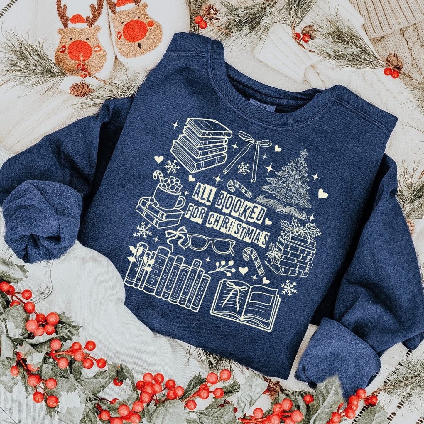 sweatshirt with a white holiday book-themed design and the phrase ‘All Booked for Christmas,’ styled flat lay with festive decor including pine branches, red berries, and cozy reindeer slippers.