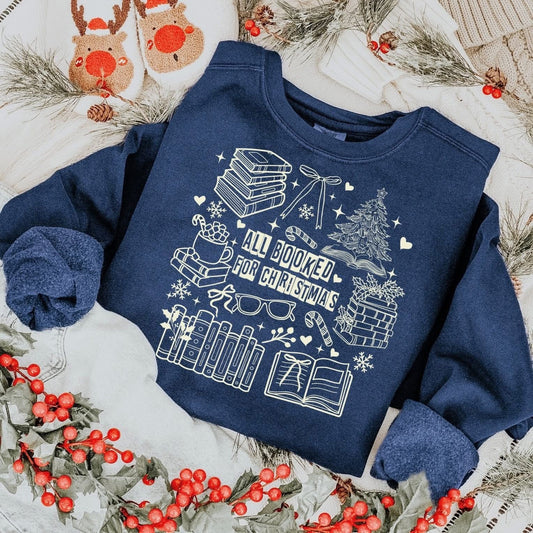 sweatshirt with a white holiday book-themed design and the phrase ‘All Booked for Christmas,’ styled flat lay with festive decor including pine branches, red berries, and cozy reindeer slippers.