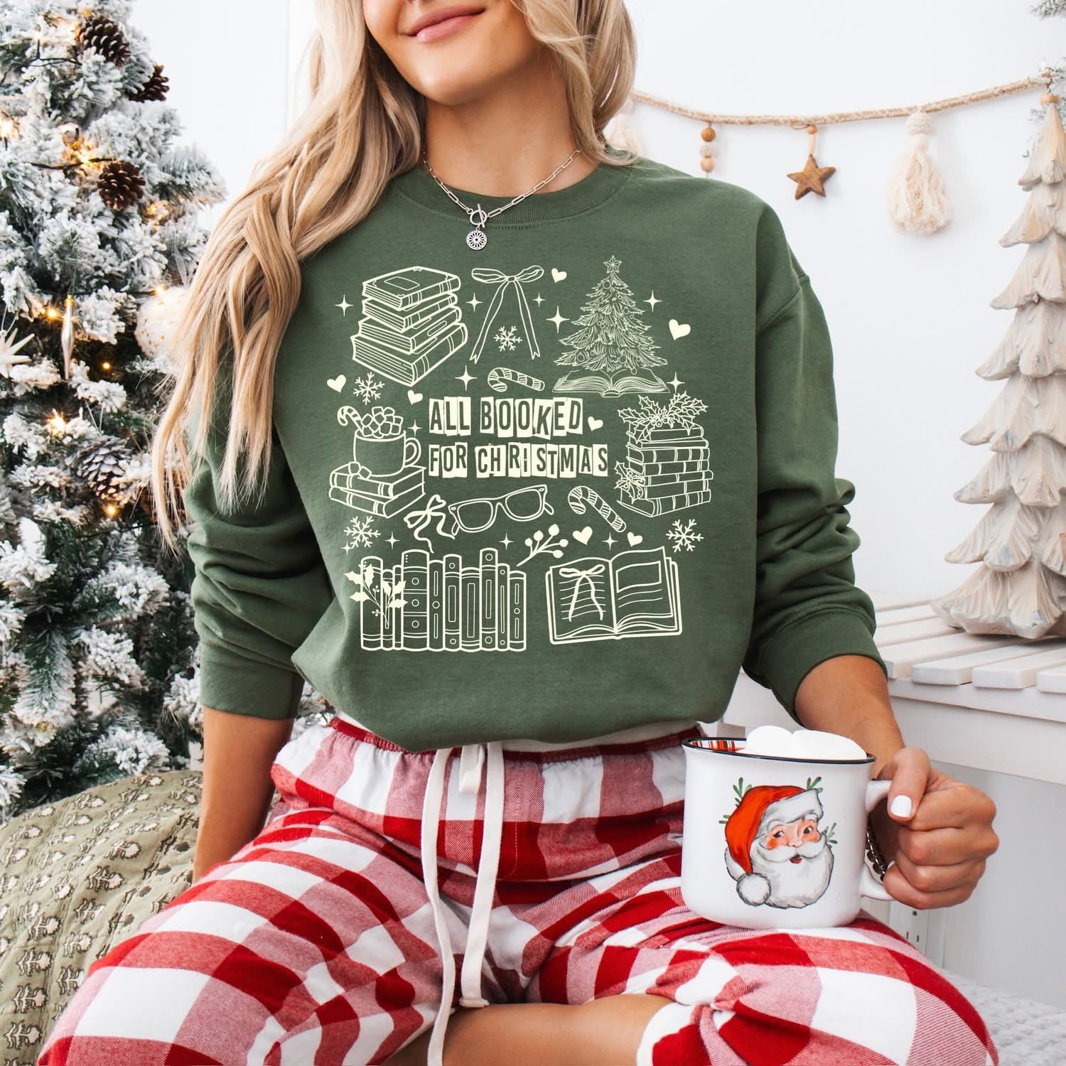 sweatshirt with a white holiday book-themed design and the phrase ‘All Booked for Christmas,’ styled flat lay with festive decor including pine branches, red berries, and cozy reindeer slippers.