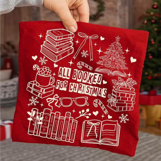 sweatshirt with a white holiday book-themed design and the phrase ‘All Booked for Christmas,’ styled flat lay with festive decor including pine branches, red berries, and cozy reindeer slippers.